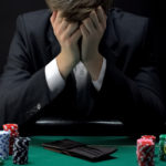 A person in a suit sitting at a poker table with their head in their hands, looking distressed, surrounded by stacks of poker chips and a wallet